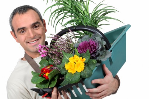 A man smiling and wearing gardening attire holds a green plastic watering can filled with a variety of colourful flowers and lush green foliage, set outdoors in a garden with a well-maintained lawn and a background of trees and shrubs. The scene suggests professional gardening or plant care, with vibrant blooms including yellow, purple, and red flowers contrasted against the green leaves. The background includes a neatly mowed lawn area, and the overall environment appears bright and clear, indicating good weather. This image reflects gardening services such as planting, flower bed maintenance, or general outdoor yard care, suitable for a professional London area garden like Honor Oak in the postcode area SE23, aligning with the services offered by Hedge Trimming Honor Oak.